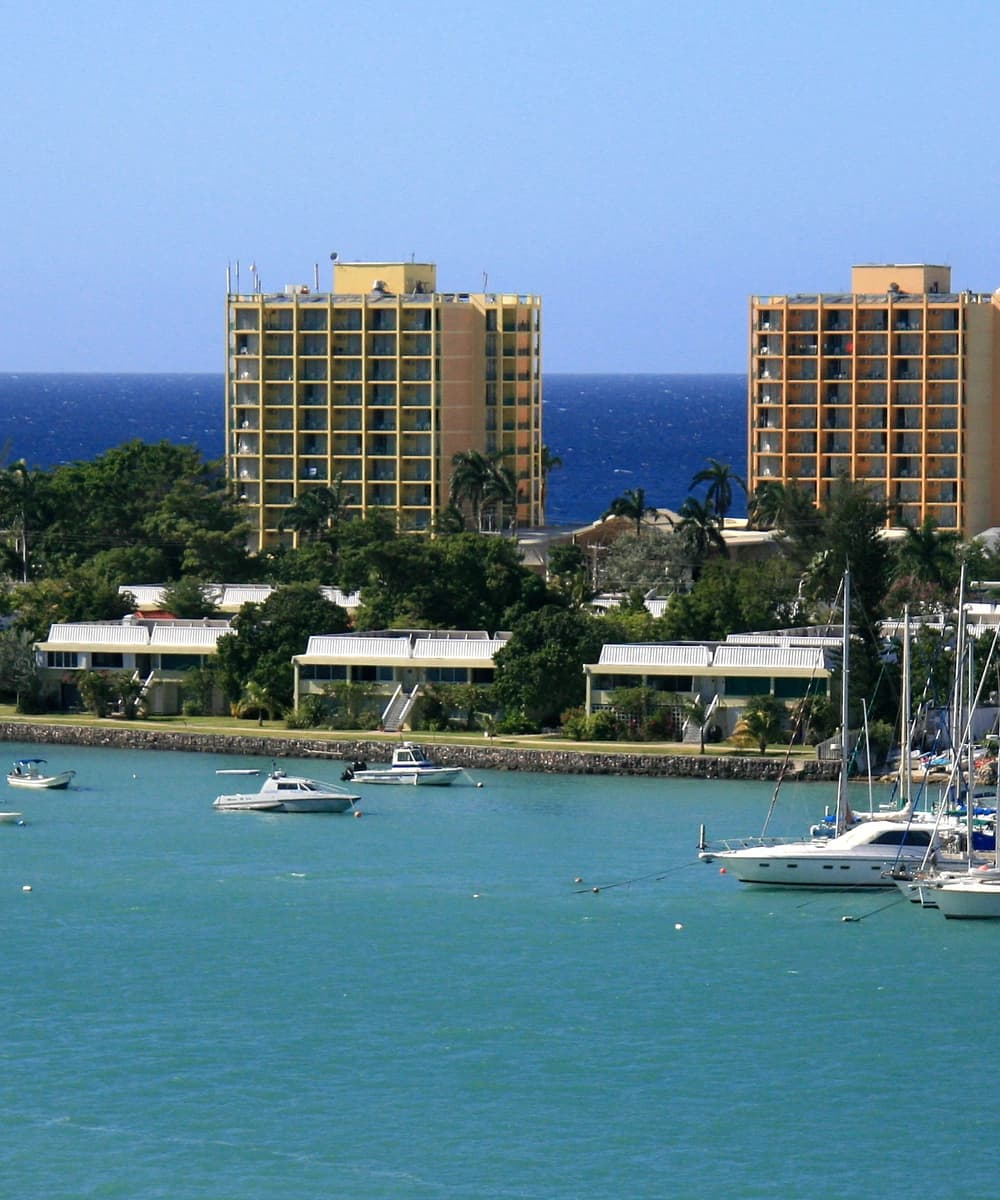 Montego Bay waterfront with yachts moored in turquoise marina water, low buildings and palms along the shore, and two towers against the open sea beyond