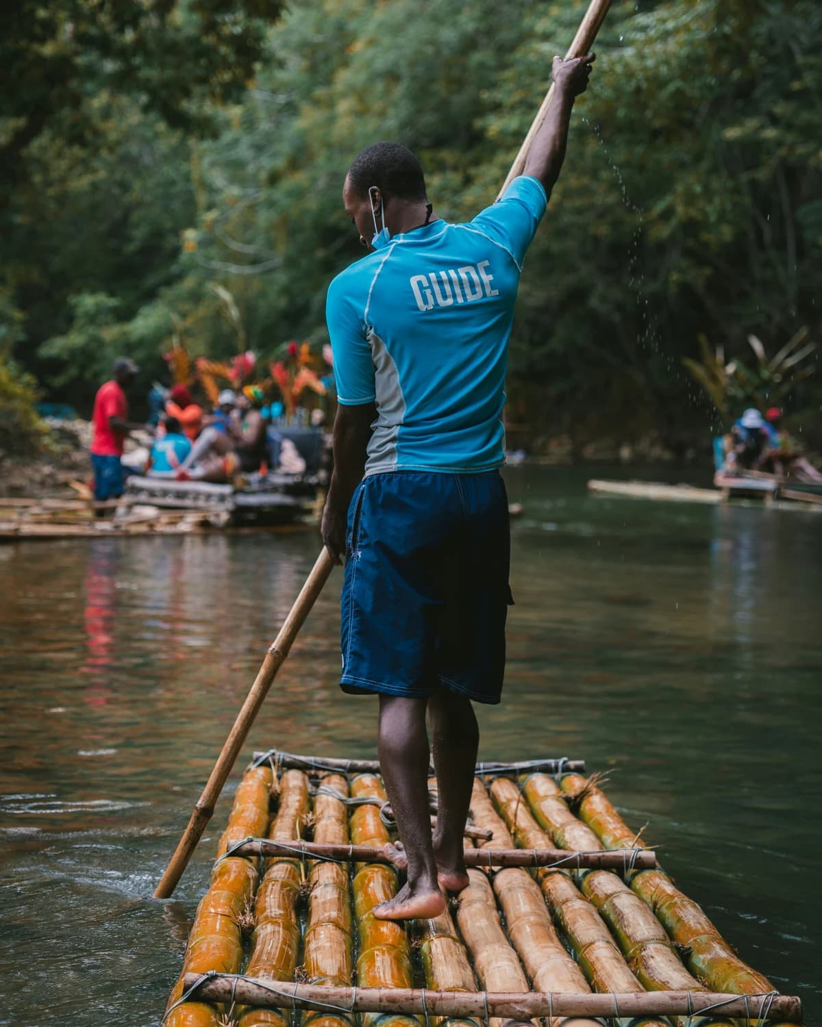 A raft captain in a blue guide shirt poling a bamboo raft along the Martha Brae, seen from behind with the pole raised and water running off it, green river and dense vegetation around him