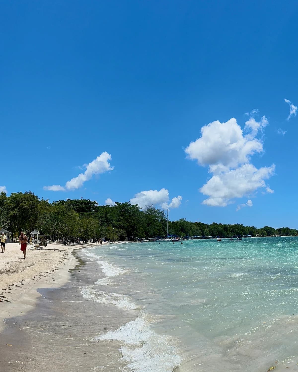 Seven Mile Beach in Negril, with pale sand in the foreground, a line of surf running along the shore, turquoise shallows opening into deeper blue water, and trees and low buildings along the coastline behind